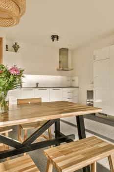 a dining table and bench in a kitchen with white cupboards behind it, there is a vase of flowers on the table