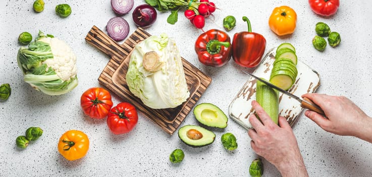 Fresh vegetables and male hands cutting zucchini with knife on wooden cutting board on white kitchen table top view. Cooking vegetarian meal from healthy ingredients, diet food and nutrition concept