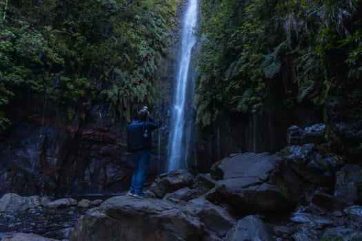 A tourist taking picture of the majestic beauty of nature, surrounded by a lush landscape with rock and water cascading down a waterfall.