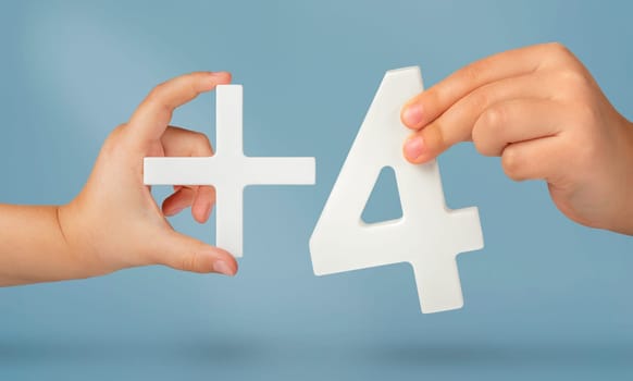 The number four and the plus symbol in the hands of a child on a blue background. White number 4 plus close up. Add three concept