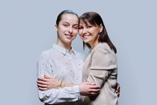 Portrait of middle aged mother and teenage daughter embracing together on grey studio background. Smiling happy mom and girl looking at camera. Family, lifestyle, relationship, mother's day concept