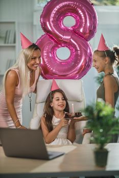 Beautiful young mom with her cute daughters have video call with friends while birthday celebration at home during pandemic isolation.