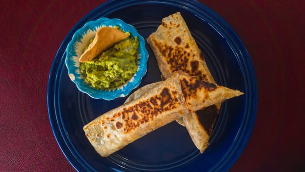 Mexican breakfast burrito. Beef machaca and egg wrapped in a fresh flour tortilla with homemade guacamole and tortilla chip. On red background and blue plate.