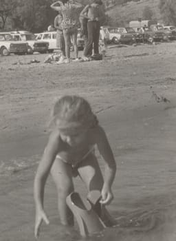 THE CZECHOSLOVAK SOCIALIST REPUBLIC - AUGUST 1977: Vintage photo shows a small girl plays in the water with swimming fins. Holidays - vacation theme. Retro black and white photography. Circa 1980.