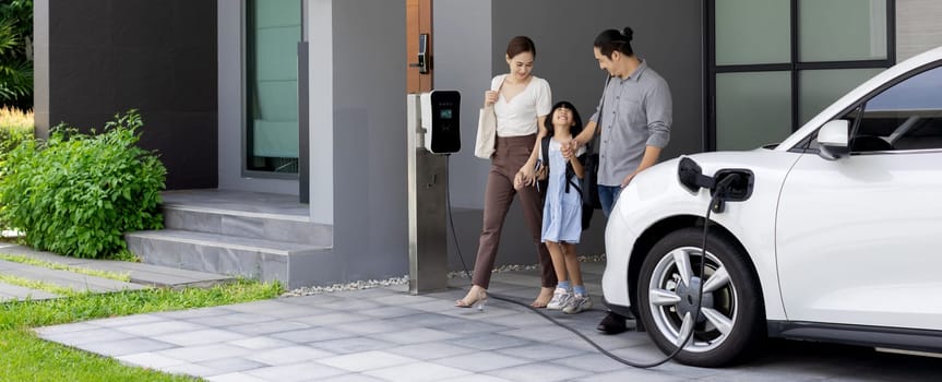 Progressive young parents and daughter with electric vehicle and home charging station. Green and clean energy from electric vehicles for healthy environment. Eco power from renewable source at home.