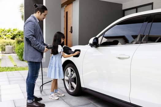 Progressive father and daughter plugs EV charger from home charging station to electric vehicle. Future eco-friendly car with EV cars powered by renewable source of clean energy.