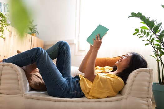 Happy young Asian woman reading a book laughing lying down on couch at home. Lifestyle concept.