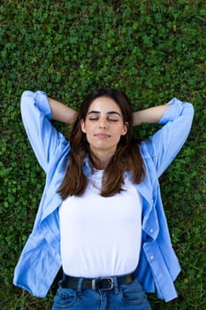 Top view vertical portrait of serene, relaxed young brunette woman lying down on grass with eyes closed. Wellbeing and lifestyle concept.