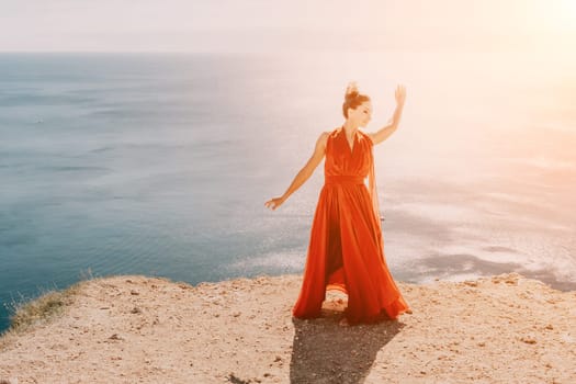 Side view a Young beautiful sensual woman in a red long dress posing on a rock high above the sea during sunrise. Girl on the nature on blue sky background. Fashion photo.