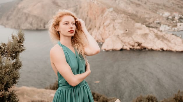 Side view a Young beautiful sensual woman in a mint long dress posing on a volcanic rock high above the sea during sunset. Girl on the nature on overcast sky background. Fashion photo