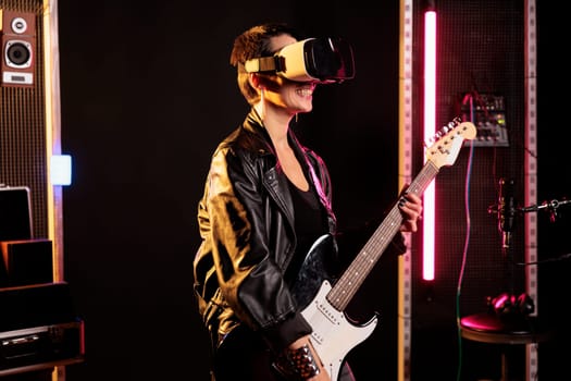 Rebel rockstar playing heavy metal song using electric guitar while wearing vr goggles to have rock concert simultation, enjoying experience in music studio. Woman musician working at new grunge album