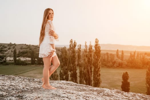 Romantic beautiful bride in white dress posing with sea and mountains in background. Stylish bride standing back on beautiful landscape of sea and mountains on sunset