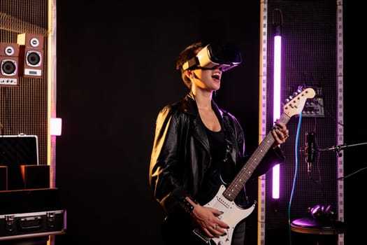 Rockstar with vr goggles enjoying rock concert simulation while playing heavy metal song at electric guitar in music studio. Woman musician performing grunge album using electricinstrument