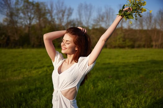 a beautiful, happy woman in a light dress stands in a field raising her hands high holding a bouquet of flowers. High quality photo