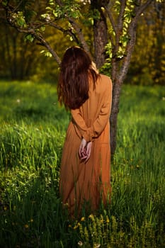 a sweet, attractive woman with long red hair stands in the countryside near a flowering tree in a long orange dress facing the camera with her hands folded on her back. High quality photo