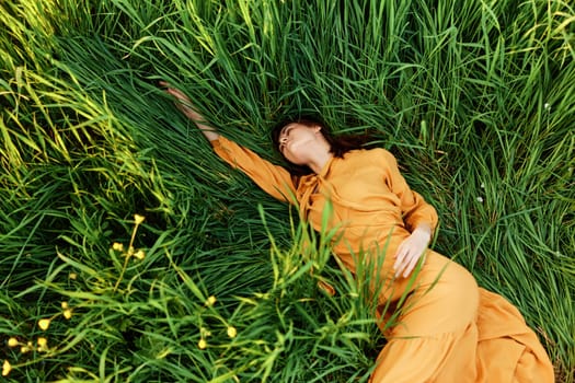 a relaxed woman enjoys summer lying in the tall green grass in a long orange dress stretched out stretching her arm forward. High quality photo