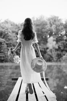 a woman in a long light dress stands on a pier by a lake in the forest with a basket and a wicker hat. monochrome photography. High quality photo