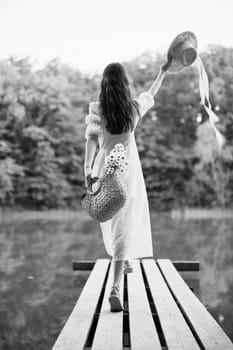 a woman in a long light dress stands on a pier by a lake in the forest with a basket and a wicker hat. monochrome photography. High quality photo