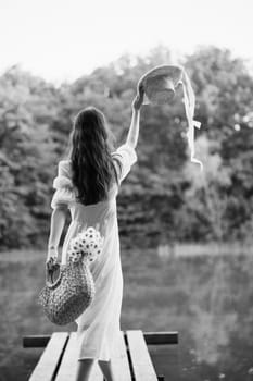 a woman with long hair stands with her back to the camera with a wicker hat in her hands on the shore of the lake. monochrome photography. High quality photo