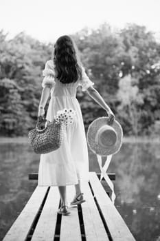 a woman in a long light dress stands on a pier by a lake in the forest with a basket and a wicker hat. monochrome photography. High quality photo