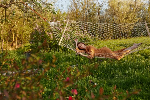 a happy woman in a long orange dress is relaxing in nature lying in a mesh hammock enjoying the peace and tranquility in the country surrounded by green foliage. High quality photo