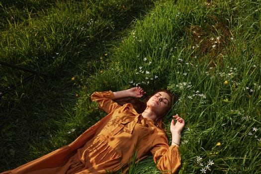 a happy, relaxed woman, resting lying in the green grass, in a long orange dress, with her eyes closed and a pleasant smile on her face, holding her hands near her face, enjoying harmony with nature and recuperating. High quality photo
