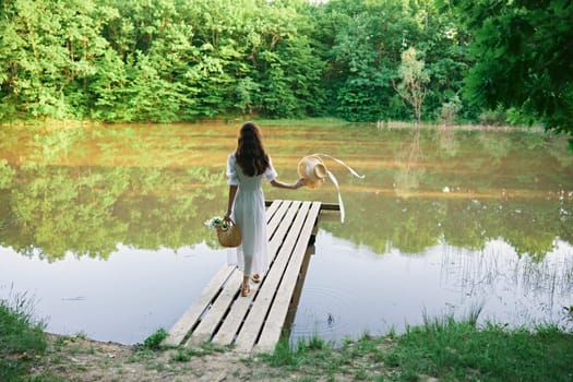 a woman in a long summer dress stands with her back to the camera on a pier near a lake in the forest with a wicker hat and a basket. High quality photo