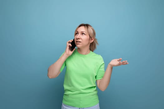 doubtful attractive blond girl holding a smartphone in her hand on a blue background with copy space.