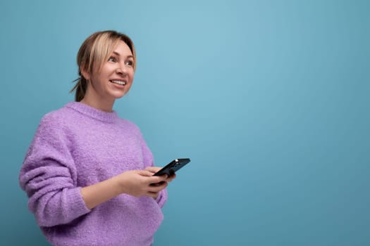 smiling blond young woman in a purple hoodie with a smartphone in her hands on a blue background with copy space.