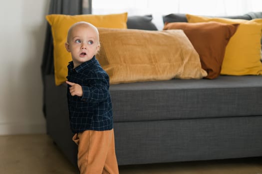 Toddler boy laughing having fun standing near sofa in living room at home. Adorable baby making first steps alone. Happy childhood and child care