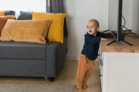 Toddler boy laughing having fun standing near sofa in living room at home. Adorable baby making first steps alone. Happy childhood and child care