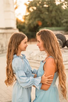 Portrait of a mother and daughter in blue dresses with flowing long hair against the backdrop of a sunset and a white building. They look at each other. Family stories on the weekend.