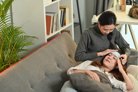 Shot of caring Asian woman taking care of sick girlfriend with fever on couch at home. LGBT, equal rights and love concept.