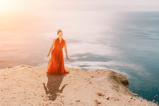 Side view a Young beautiful sensual woman in a red long dress posing on a rock high above the sea during sunrise. Girl on the nature on blue sky background. Fashion photo.