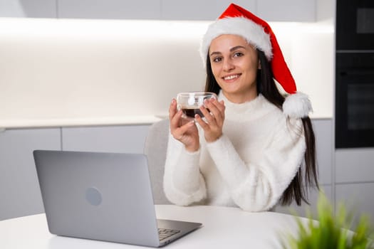 Portrait of a happy beautiful woman in a Santa Claus hat holding a cup of coffee while working using a notebook at home.