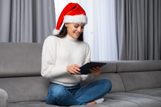 A happy young woman dressed in a red Santa Claus hat sitting on the sofa and using a tablet. Congratulation on Merry Christmas and Happy New Year.