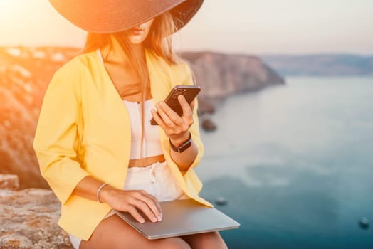 Successful business woman in yellow hat working on laptop by the sea. Pretty lady typing on computer at summer day outdoors. Freelance, travel and holidays concept.