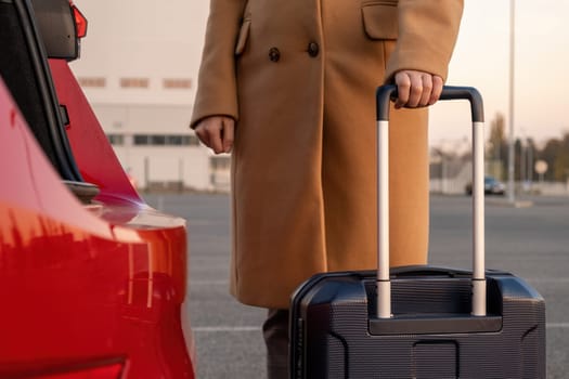 A woman in a brown coat stands with a suitcase near a red car. Shuttle from an airport concept