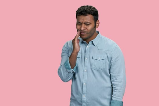 Sad unhealthy indian or arabian guy touching cheek with hand with painful expression. Toothache or dental illness. Isolated on pink.