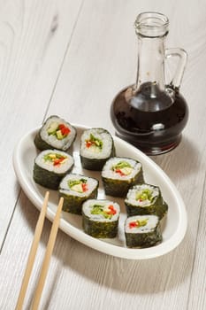 Sushi rolls with rice, pieces of avocado, cucumber, red bell pepper and lettuce leaves on ceramic plate, chopsticks, glass bottle with soy sauce on wooden desk