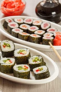 Different sushi rolls with rice and seaweed sheets on ceramic plate, glass bottle with soy sauce and pickled ginger in a bowl