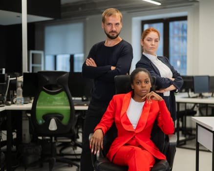 Caucasian red-haired woman, bearded Caucasian man stand behind a seated African American young woman in the office