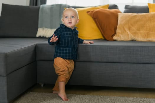 Toddler boy laughing having fun standing near sofa in living room at home. Adorable baby making first steps alone. Happy childhood and child care