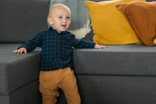 Toddler boy laughing having fun standing near sofa in living room at home. Adorable baby making first steps alone. Happy childhood and child care