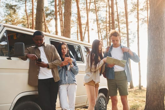 Standing near the car. Group of young people is traveling together in the forest at daytime.