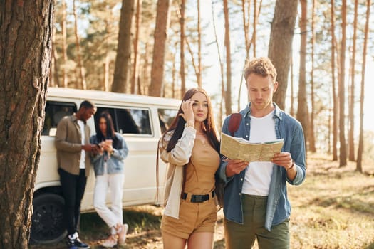 Standing near the car. Group of young people is traveling together in the forest at daytime.