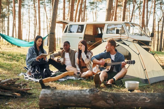 Man plays guitar. Group of young people is traveling together in the forest at daytime.