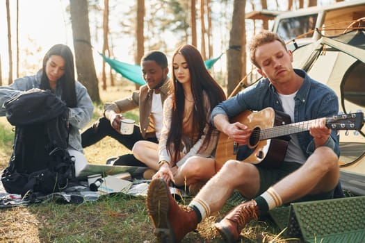 Man plays guitar. Group of young people is traveling together in the forest at daytime.