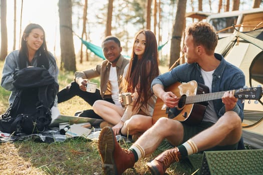 Man plays guitar. Group of young people is traveling together in the forest at daytime.