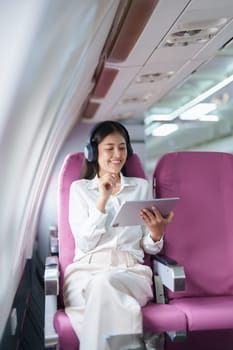 Young Asian attractive business woman passenger sitting on business class luxury plane while wearing wireless headphone for relax and working with tablet during the flight.